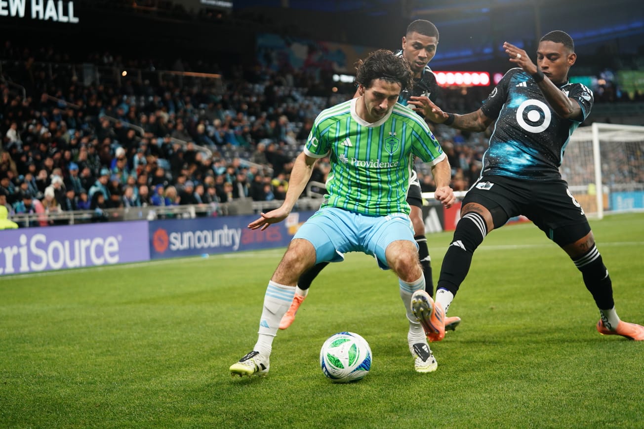 Paul Rothrock, wearing a green Sounders jersey and blue shorts, defends the ball from two defenders.